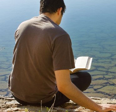 Photo of a young man reading the Bible by a lake. The Bible is where God shows that He has a plan and is in control of all things, but also that He has allowed people to have free will, which has resulted in all sorts of problems.